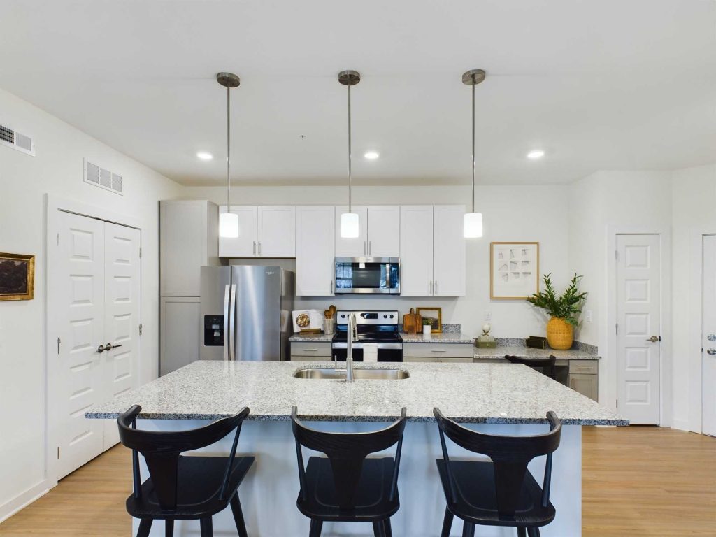 Apartments in Thompson’s Station Modern kitchen with a granite island, three black chairs, stainless steel appliances, and pendant lighting. Light wood flooring and white cabinets complete the space. Sanctuary Bluff Apartments in Thompson’s Station