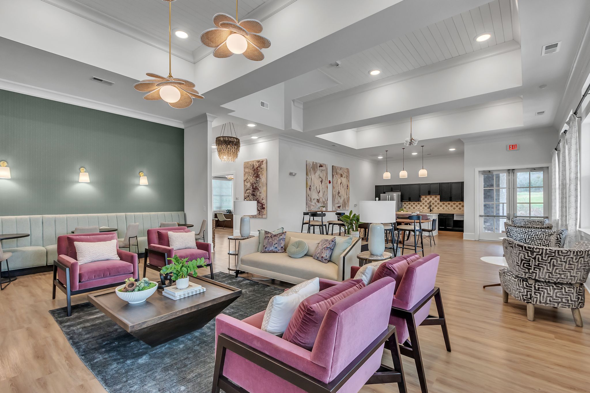 Apartments in Thompson’s Station A modern living room with pink chairs, a beige sofa, and patterned armchairs. The room has a green accent wall, wooden flooring, and pendant lights. A kitchen is visible in the background. Sanctuary Bluff Apartments in Thompson’s Station