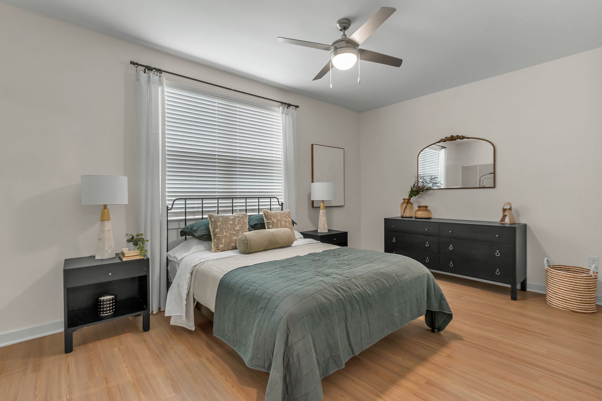 Apartments in Thompson’s Station A bedroom with a bed, green bedding, two nightstands with lamps, a dresser with a mirror, and a ceiling fan. The room has wooden floors and a window with blinds. Sanctuary Bluff Apartments in Thompson’s Station