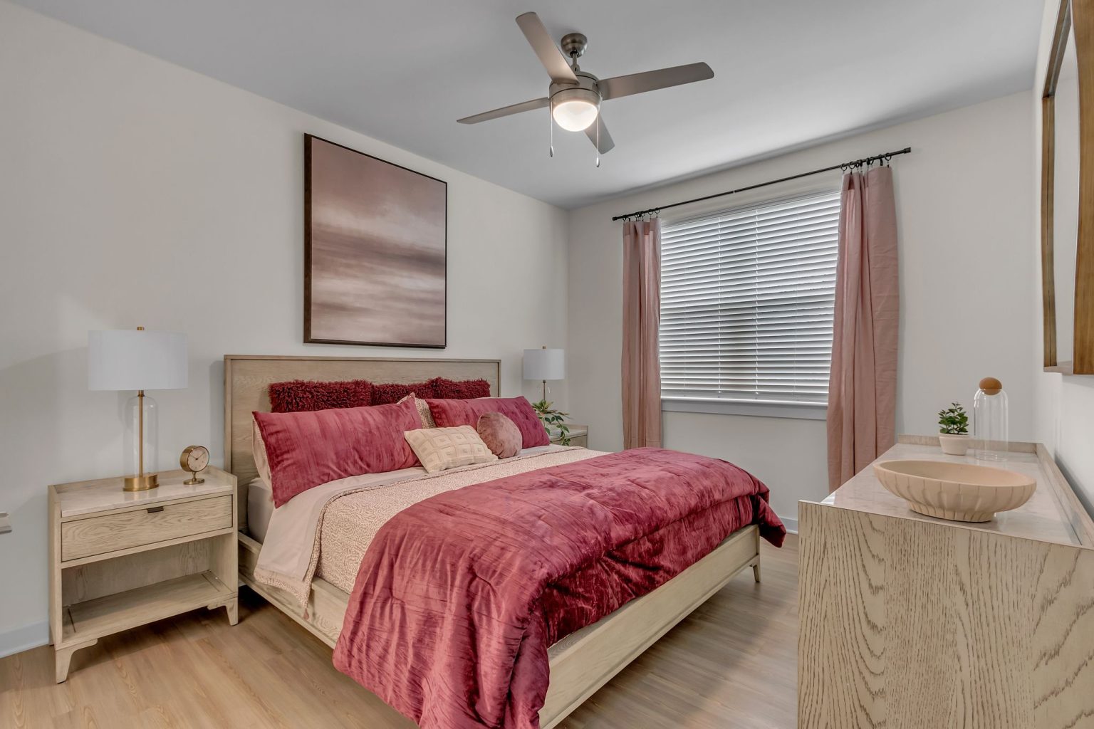 Apartments in Thompson’s Station Bedroom with a bed featuring pink bedding, two side tables with lamps, a ceiling fan, a large window with pink curtains, and a decorative painting above the headboard. Sanctuary Bluff Apartments in Thompson’s Station
