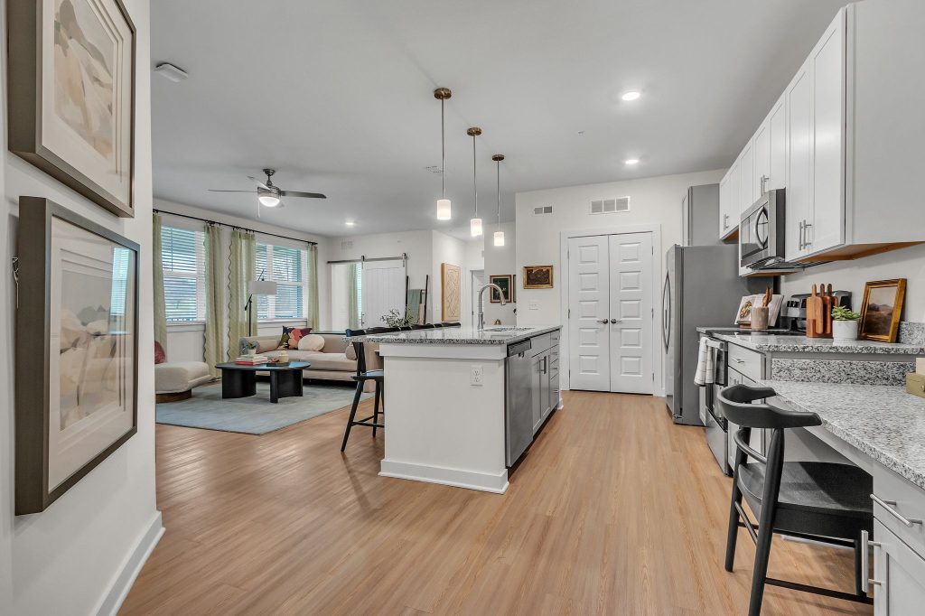 Apartments in Thompson’s Station Modern kitchen and living area with white cabinets, stainless steel appliances, and wooden flooring. There's a central island with bar stools and a sofa in the adjacent living space. Sanctuary Bluff Apartments in Thompson’s Station