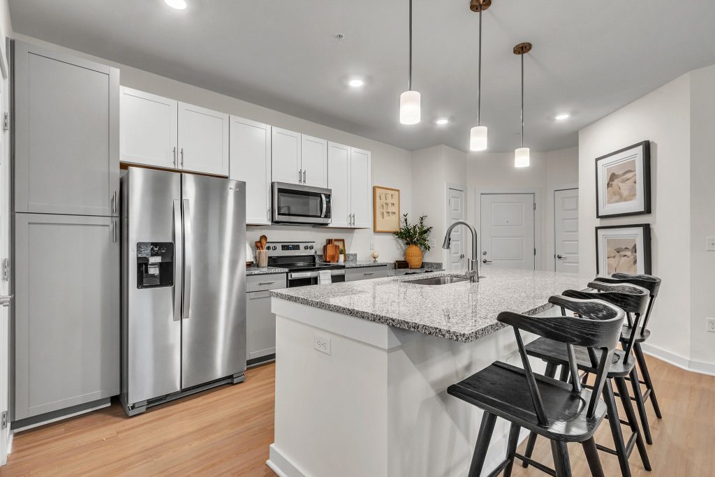 Apartments in Thompson’s Station Modern kitchen with stainless steel appliances, white cabinets, and a large granite island. Three black stools are placed at the island under pendant lights. Wood flooring throughout. Sanctuary Bluff Apartments in Thompson’s Station