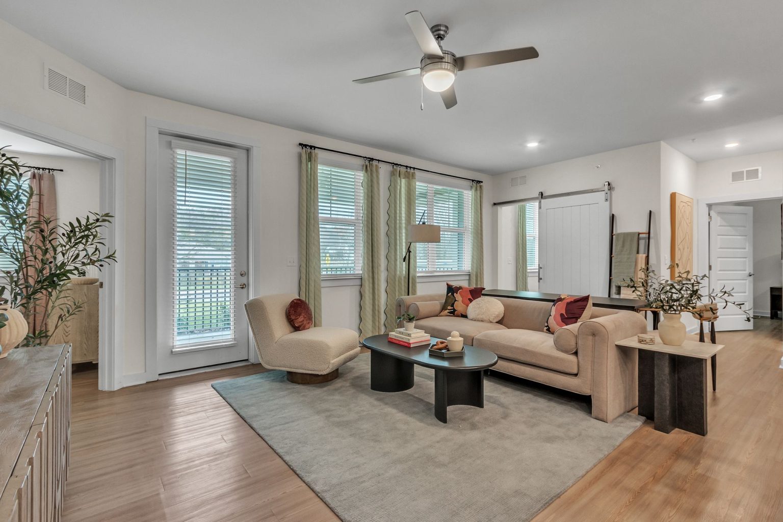 Apartments in Thompson’s Station A modern living room with a beige sofa, round chair, coffee table, plants, and ceiling fan. Wooden flooring and a rug complement the large windows with sheer curtains. Sanctuary Bluff Apartments in Thompson’s Station