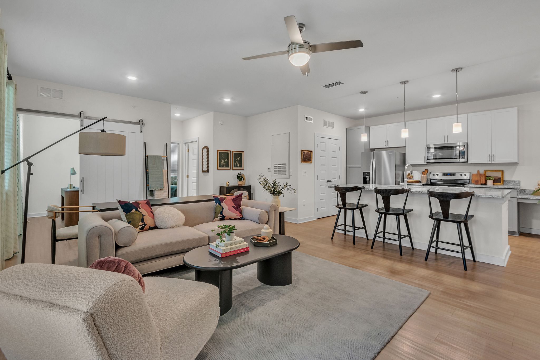 Apartments in Thompson’s Station Modern open-concept living room and kitchen with neutral-toned furniture, wooden floor, artistic decor, and ceiling fan. Four stools at a kitchen island under pendant lights. Sanctuary Bluff Apartments in Thompson’s Station