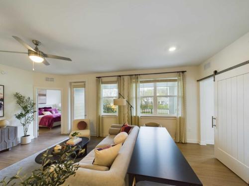 Apartments in Thompson’s Station A modern living room with a ceiling fan, beige sofa, and black table. A view into a bedroom with pink bedding is visible through an open doorway. Large windows offer natural light. Sanctuary Bluff Apartments in Thompson’s Station