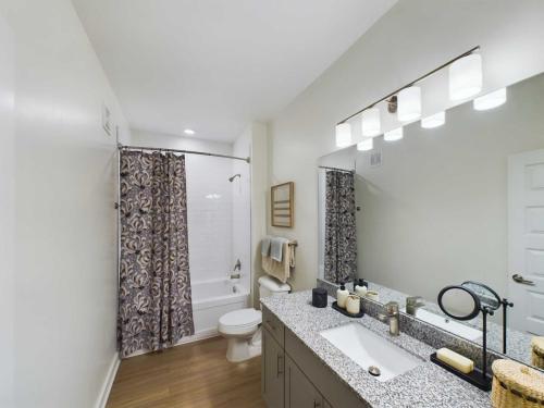 Apartments in Thompson’s Station Modern bathroom with a shower-tub combo, floral curtain, granite countertop, large mirror, and vanity lighting; wooden floor and light-colored walls create a clean, minimalistic look. Sanctuary Bluff Apartments in Thompson’s Station