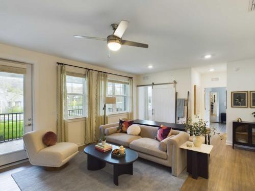 Apartments in Thompson’s Station Living room with a beige sofa, round chair, black coffee table, ceiling fan, and large windows. Decor includes books, plants, and wall art. Light wooden floors and a sliding barn door are visible. Sanctuary Bluff Apartments in Thompson’s Station