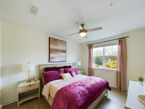 Apartments in Thompson’s Station A bedroom with a bed featuring magenta bedding, two bedside tables with lamps, a ceiling fan, and a large window with pink curtains, overlooking a construction site. Sanctuary Bluff Apartments in Thompson’s Station