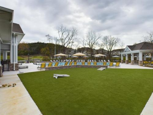 Apartments in Thompson’s Station Outdoor seating area with lounge chairs and umbrellas near a pool, surrounded by trees and buildings, under a cloudy sky. There's a grassy area with a cornhole game in the foreground. Sanctuary Bluff Apartments in Thompson’s Station