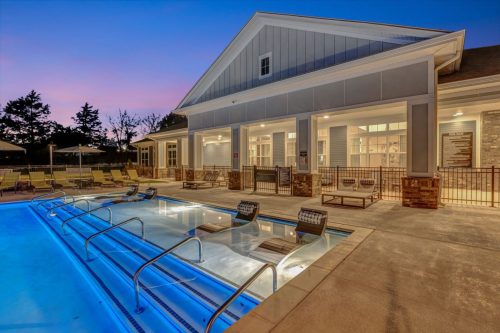 Apartments in Thompson’s Station Outdoor swimming pool area with lounge chairs partially submerged in the water, adjacent to a lit, contemporary building at dusk. Sanctuary Bluff Apartments in Thompson’s Station