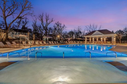 Apartments in Thompson’s Station A lit swimming pool area at dusk, surrounded by lounge chairs and a pavilion on the right side, with trees and houses in the background. Sanctuary Bluff Apartments in Thompson’s Station